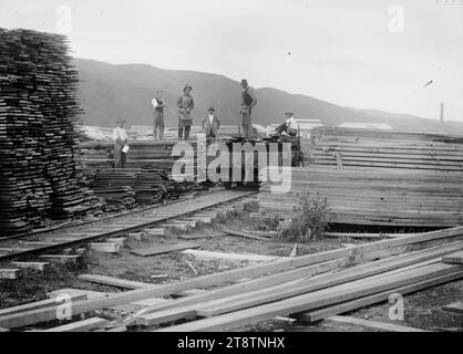 Holz gestapelt auf einem Sägewerk, Schnittholz neben einer Eisenbahnstrecke und auf einem Drehgestell in einem Holzhof. Ein Schornstein und Gebäude sind in der Ferne zu sehen. An einem unbekannten Ort, ca. 1920 Eine nicht identifizierte Gruppe von Sägewerkarbeitern steht auf den gestapelten Sägeplanken Stockfoto