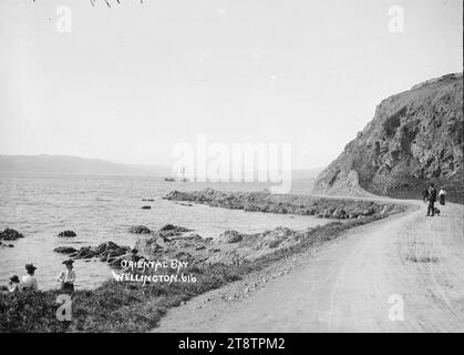 Oriental Bay, Wellington, Neuseeland, Blick auf Lambton Harbour von der Oriental Parade aus in Richtung Nordosten in Richtung Petone mit Somes Island in der Ferne. Ein Fischerboot ? Geht auf den Weg ins Meer. Zwei Frauen und ein Junge stehen auf den Felsen am Ufer im Vordergrund links. Mehrere Leute sind auf der Straße, einer fährt mit dem Fahrrad. Foto zwischen 1910 und 1930 Stockfoto