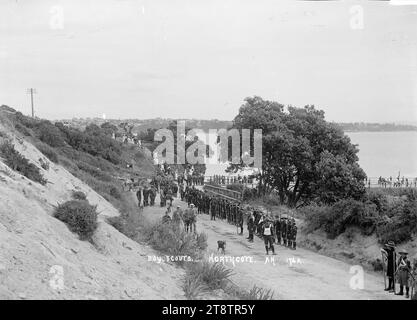 Pfadfinder in Northcote, Auckland, Neuseeland. Pfadfinder stehen auf der Queen Street an der Northcote Wharf. Sie sind auf der Parade für die Präsentation im flagstaff am Stokes Point im Jahr 1908 zur Gründung des Borough. Die Späher halten Stab und einige tragen Fahnen. Männer und Frauen laufen die Stufen von einer Straße oben hinunter und die Leute stehen neben einem Pferdebus in der Nähe des Kais. Auckland, Neuseeland, kann man in der Ferne über den Waitemata Harbour sehen Stockfoto