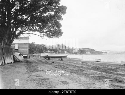 Boote in Hall's Beach, Northcote, Auckland, Neuseeland, Blick auf Hall's Beach mit Blick auf die Northcote Wharf mit Auckland, Neuseeland in der Ferne. Ein Bootsschuppen mit Schild oben (Bailey & Tyer - nicht sichtbar) befindet sich auf der linken Seite. Vor dem Bootsschuppen in einer Jacht, und die Bootsrampe ist einfach dahinter sichtbar. Zwei Schlauchboote sind am Wasserrand verankert. Häuser können auf der Klippe mit Blick auf Little Shoal Bay, CA 1910, besichtigt werden Stockfoto