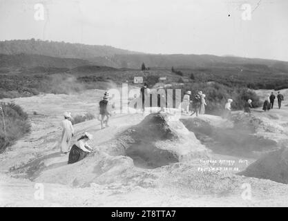 Tikitere, Rotorua County, Blick auf das Thermalgebiet von Tikitere, bei dem eine Gruppe von Besuchern durch die Gegend geführt wird. Eine Frau im Vordergrund kniet nieder und schaut auf die Erdkruste. Im Jahr 1908 gibt es zwei Häuser in der Mitteldistanz Stockfoto