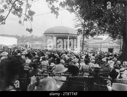 Menschen, die den Empfang der Massenbands hören, Auckland, Neuseeland-Ausstellung, Foto von Menschen, die auf Stühlen unter Bäumen sitzen und den Empfang eines Mannes hören, der in einer Rotunde steht. Hinter ihm sitzen andere Würdenträger. Rechts von der Rotunde befindet sich das Technische College-Gericht. Foto aufgenommen bei der Auckland, Neuseeland Ausstellung in Auckland, Neuseeland, 11. Februar 1914 Stockfoto