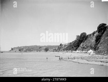 Blick auf Orakei Beach, Auckland, Neuseeland, Blick auf Orakei Beach (jetzt Teil des Tamaki Drive) mit Blick nach Osten in Richtung Bastion Point. Zelte werden am Fuß der Klippe errichtet und die Menschen waten im Meer, laufen am Strand entlang und stehen am felsigen Ufer. Häuser in der Biddicks Bay sind in der Entfernung ca. 1910 zu sehen Stockfoto