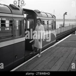 1950er Jahre, historisch, eine gut gekleidete Dame, die auf einem hölzernen Bahnsteig steht und gerade an Bord eines elektrischen Zuges am Pier in Southend on Sea geht. Im Eisenbahnwagen eine Nachricht für die Southend Regatta. Diese Pier-Eisenbahnwaggons wurden 1949 eingeführt und bis Ende der 1970er Jahre betrieben, als ein kleinerer Dieselzug eröffnet wurde. Stockfoto