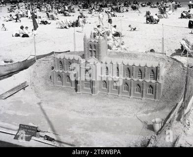 1950er Jahre, historisch, am Strand, Menschen auf Liegestühlen und ein Sandmodell von Bath Abbey, England, Großbritannien. Die Überschrift besagt: „Dieses Modell besteht nur aus Sand und Meerwasser“. Die 1611 eröffnete Bath Abbey in Somerset, offiziell bekannt als Abteikirche St. Peter und St. Paul, ist ein im gotischen Stil erbautes Benediktinerkloster. Stockfoto