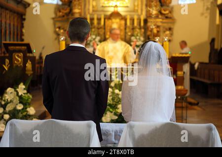 Ein junges Paar steht vor dem Altar während einer Hochzeitszeremonie in einer katholischen Kirche. Ein Priester feiert die Messe im Hintergrund. Stockfoto