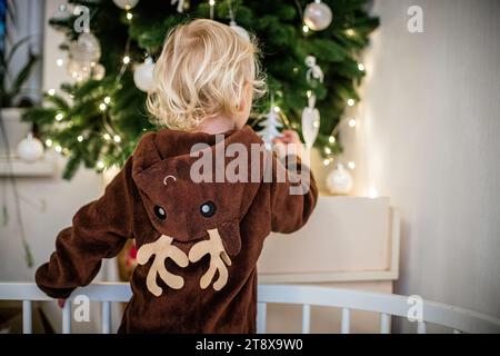 Kleines Mädchen im Rentierkostüm, das den Weihnachtsbaum schmückt, während es in der Krippe steht Stockfoto