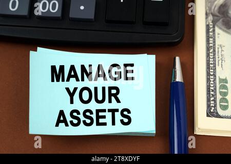 Geschäftskonzept. Auf einer braunen Oberfläche liegen ein Taschenrechner, Dollar, Stifte und Aufkleber mit der Inschrift - verwalten Sie Ihre Assets Stockfoto