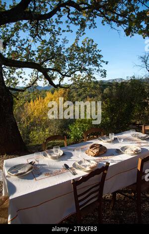 Une table à Manger dressée, avec assiettes, couverts verres et miche de Pain, sur une Terrasse à l'heure du souper, avec en arrière Plan les collines Stockfoto