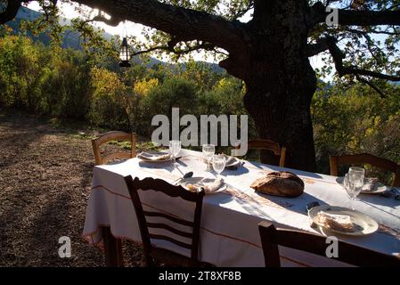 Une table à Manger dressée, avec assiettes, couverts verres et miche de Pain, sur une Terrasse à l'heure du souper, avec en arrière Plan les collines Stockfoto