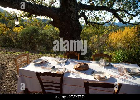 Une table à Manger dressée, avec assiettes, couverts verres et miche de Pain, sur une Terrasse à l'heure du souper, avec en arrière Plan les collines Stockfoto