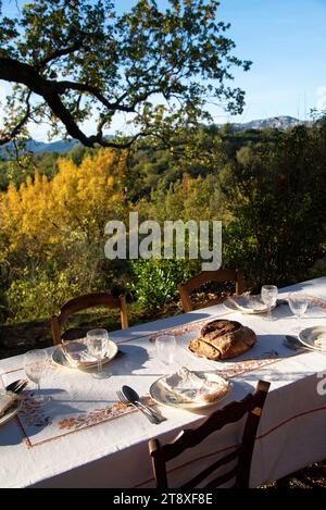 Une table à Manger dressée, avec assiettes, couverts verres et miche de Pain, sur une Terrasse à l'heure du souper, avec en arrière Plan les collines Stockfoto