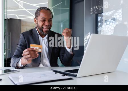 Glücklicher junger afroamerikanischer Mann, der im Büro vor dem Laptop sitzt und glücklich ist, Kreditkarte hält und Sieg zeigt Ja-Geste. Stockfoto