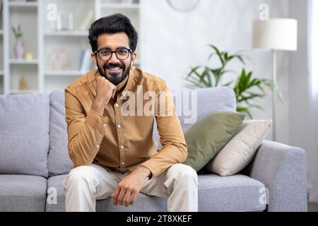 Porträt eines jungen Indianers, der zu Hause auf dem Sofa sitzt, seine Hand auf dem Kinn legt, selbstbewusst und lächelnd in die Kamera schaut. Stockfoto