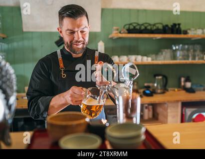 Ein lächelnder, stilvoller Bärenbarmann in schwarzer Uniform mit einer Schürze, der frisches Lagerbier in einen Glasbecher an der Bar zapft. Erfolgreiche Leute, Bierkonzern Stockfoto