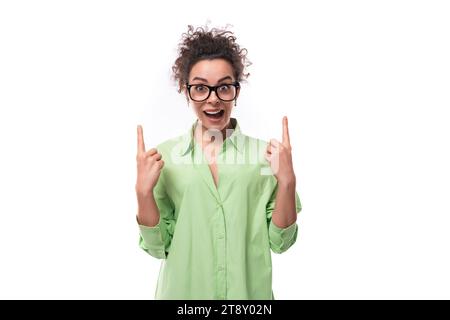 Hübsche junge, brünette lockige Frau in Brille in einem grünen Hemd zeigt ihre Hand zur Seite auf einem weißen Hintergrund mit Kopierraum Stockfoto