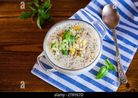 Kalte Suppe mit Gurken, Eiern, Gemüse, Joghurt und frischen Kräutern in einer Glasschale auf einem Holztisch. Stockfoto