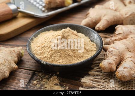 Schüssel mit Ingwerpulver und ganzen Ingwerwurzeln auf dem Küchentisch. Eine Reibe mit geriebener Ingwerwurzel im Hintergrund. Stockfoto