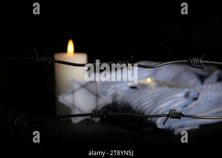 Stacheldraht mit brennenden Kerzen und Gefangenenuniform auf dunklem Hintergrund, Nahaufnahme. Internationaler Holocaust-Gedenktag Stockfoto