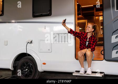 Junge Frau, die sich selbst porträtiert, während sie in der Tür eines Wohnmobil-Wohnwagens sitzt. Wohnmobilparkplätze. Stockfoto