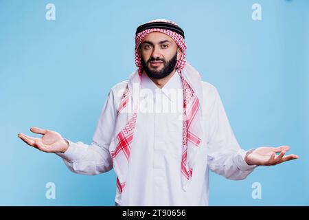 Verwirrter muslimischer Mann, der traditionelle Kleidung trägt und Schultern zuckt und mit ungewissem Ausdruck in die Kamera blickt. Arabische Person, in der sie Hände spreizt Studio-Porträt Stockfoto