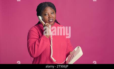 afroamerikanerin in stilvollem Outfit, die sich über das Telefon unterhalten kann. Weibliche Modebloggerin, die auf einem Festnetztelefon vor einem isolierten rosa Hintergrund spricht. Stockfoto