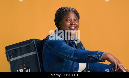 Jugendliche Kurierin, die modernes Fahrrad fährt, um Pakete effizient durch die Straßen der Stadt zu liefern. afroamerikanische Frau mit Lebensmitteltasche, die aktiv auf Abruf arbeitet. Stockfoto