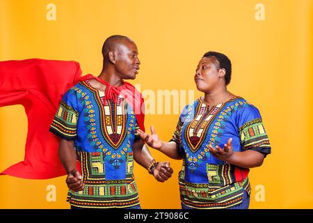 afroamerikanerin, die gestikuliert und einen Mann ansieht, der in superman cape gekleidet ist. Ein lächelnder Freund trägt einen roten Superheldenmantel und kommuniziert mit seiner Freundin im Studio Stockfoto