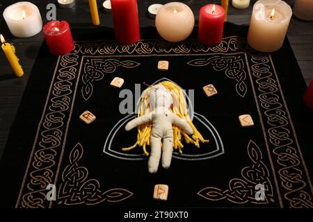 Voodoo-Puppe mit Nadeln, Runen und Kerzen auf schwarzer Matte durchbohrt. Fluchzeremonie Stockfoto