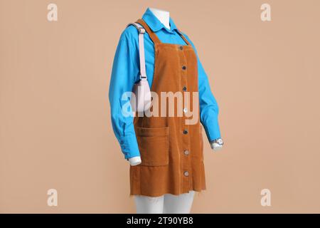 Weibliche Schaufensterpuppe mit Accessoires, die in hellblauem Hemd und orangefarbenem Pullover-Kleid auf beigem Hintergrund gekleidet sind Stockfoto