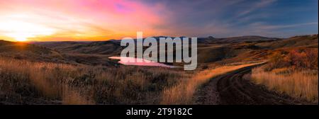 Eine Schotterstraße schlängelt sich durch die Hügel und Berge in einen pulsierenden Sonnenuntergang in diesem Panorama des Hinterlandes in der Nähe von Hayden, Colorado. Lebendiger Sonnenuntergang. Stockfoto