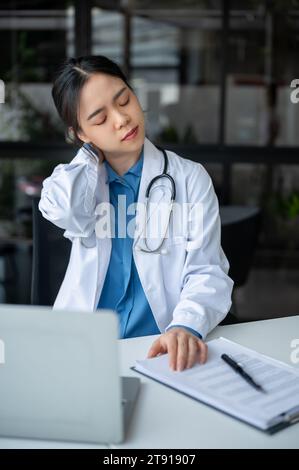 Eine müde und überarbeitete junge asiatische Ärztin massiert ihren Hals, leidet unter Nackenschmerzen und Bürosyndrom, während sie an ihrem Schreibtisch im arbeitet Stockfoto