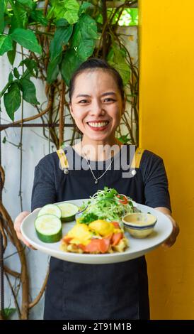 Junge vietnamesische Kellnerin, die im Restaurant einen Teller mit Eiern und Waffeln mit Räucherlachs hält Stockfoto