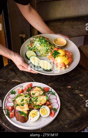 Kellnerin hält einen Teller mit Eiern, bennedict-Waffeln mit geräuchertem Lachs und serviert auf einem Tisch mit Hähnchentoast im Restaurant Stockfoto