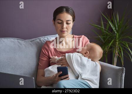 Porträt einer jungen Frau, die das schlafende Baby in den Armen hält und das Smartphone benutzt, während sie sich zu Hause auf der Couch entspannt, im Kopierraum Stockfoto