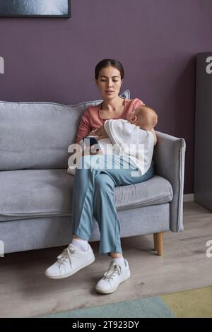 Porträt in voller Länge einer jungen Mutter, die das schlafende Baby in den Armen hält und das Smartphone benutzt, während sie sich zu Hause auf der Couch entspannt Stockfoto