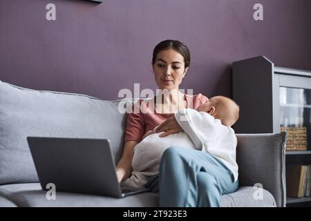 Porträt einer jungen Frau, die schlafendes Baby in den Armen hält und Laptop benutzt, während sie von zu Hause aus im Mutterschaftsurlaub arbeitet, Kopierraum Stockfoto