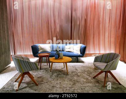 Modernes, minimalistisches Wohnzimmer mit einem eleganten blauen Sofa, zwei gestreiften Sesseln und einem hölzernen Kaffeetisch vor einem strukturierten rosafarbenen Hintergrund Stockfoto