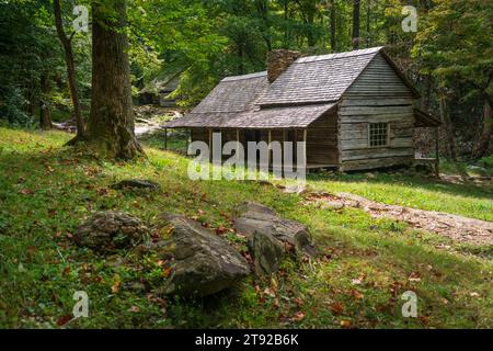 Moosige Felsen und ein Baumstamm rahmen eine entfernte graue Schindeldachhütte auf einem grasbewachsenen Ufer über einem Bach im Great Smoky Mountains National Park ein. Stockfoto