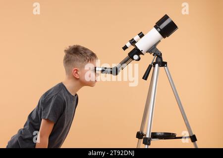 Kleiner Junge, der Sterne durch ein Teleskop auf beigefarbenem Hintergrund ansieht Stockfoto