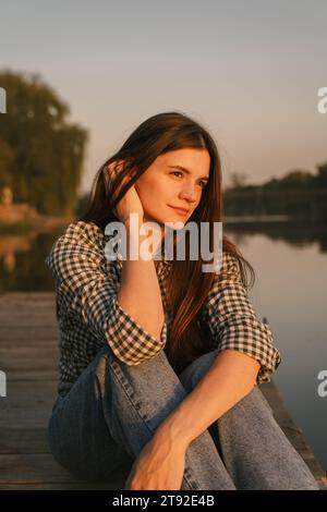 Ein Mädchen mit langen dunklen Haaren, das auf dem Flussdock sitzt. Es ist Sommer Sonnenuntergang. Das Wetter ist warm und ruhig. Stockfoto