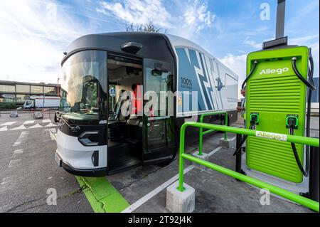 Ein Volta Zero Truck mit offener Tür steht neben einer Allego Ladestation mit einem Gebäude und einem geparkten Truck im Hintergrund vor einem klaren blauen Himmel Stockfoto