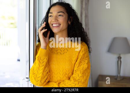 Glückliche birassische Frau in gelbem Pullover, die zu Hause auf dem Smartphone spricht. Lebensstil, Kommunikation und häusliches Leben unverändert. Stockfoto