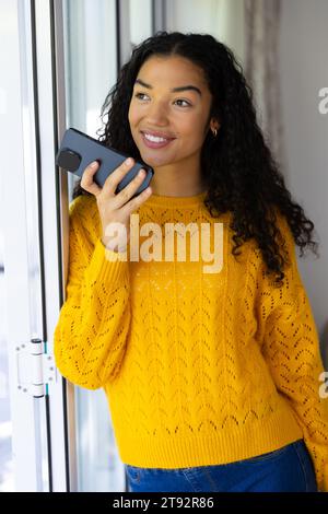 Glückliche birassische Frau in gelbem Pullover, die zu Hause auf dem Smartphone spricht Stockfoto