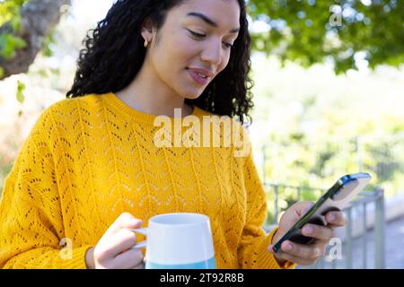 Birassische Frau in gelbem Pullover, die eine Tasse Kaffee hält und Smartphone auf der sonnigen Terrasse benutzt Stockfoto