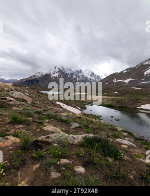 Der Pensila-See, umgeben von schneebedeckten Gipfeln, enthüllt einen ruhigen Himmel. Wildblumen zieren Wiesen und spiegeln die Harmonie der Natur im ruhigen Wasser wider. Stockfoto