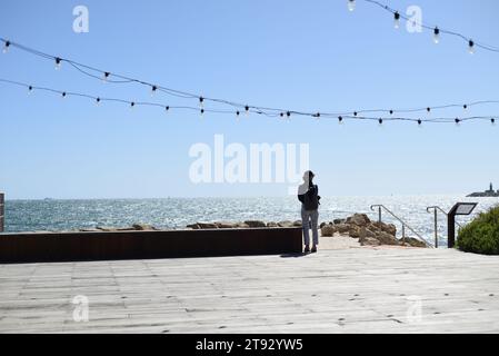 Eine Frau steht auf einer Holzterrasse mit Blick auf den Indischen Ozean. Ein leuchtend glitzernder meerblauer Himmel und Festoon Lichter, Fremantle W.A. Stockfoto