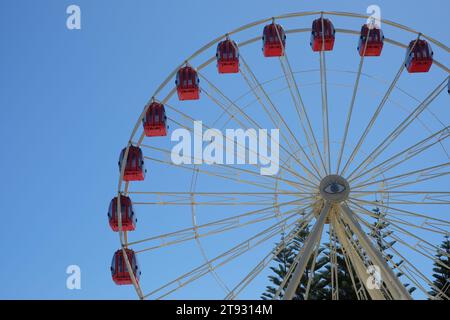 Rote Gondeln säumen den Umfang des Tourist Wheel Fremantle, eine beliebte Attraktion des Riesenrades, mit einem zentralen Augenmotiv vor einem blauen Himmel Stockfoto