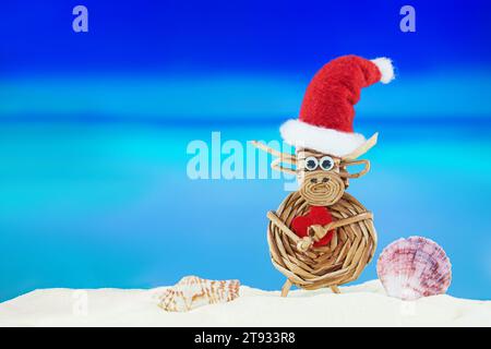 Kuh aus Spielzeuggewebe mit Weihnachtsmann-Hut mit Herz und Muscheln am Sandstrand mit Meer. Weihnachten, Neujahr, Valentinstag. Heiße Länder. Kopierbereich Stockfoto