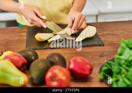 Zugeschnittene Ansicht einer vegetarischen Frau, die reife Birnen in der Nähe frischer Früchte in der Küche schneidet, pflanzliche Ernährung Stockfoto